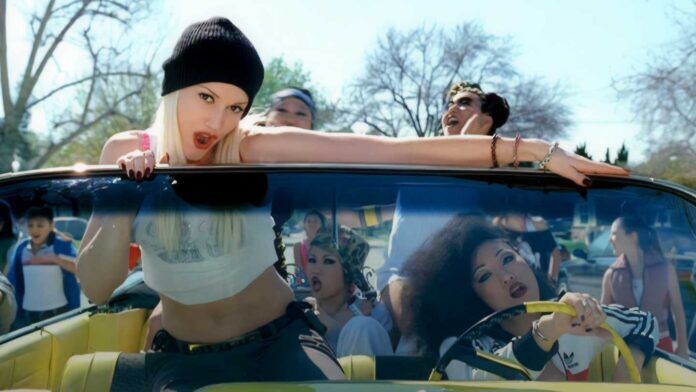 Gwen Stefani's Ain't No Hollaback Girl - A group of people in a convertible car. The front passenger, wearing a black beanie and white tank top, leans out energetically. The driver, wearing a headscarf, drives while others in the backseat chat and smile. Trees are visible in the background.