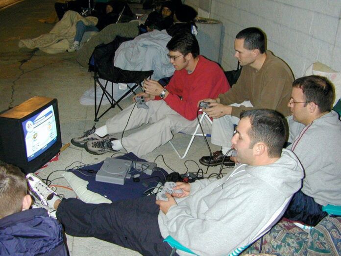 A group of people sitting on folding chairs and the ground, playing video games on a small CRT TV outside. There are portable chairs, blankets, and a PlayStation console. Other people are lying on the ground in sleeping bags in the background.