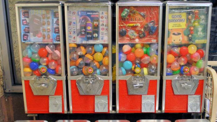 Four red and silver vending machines contain colorful plastic capsules. Each machine has a coin slot marked 50¢, and displays various toy options like mini figurines and keychains.
