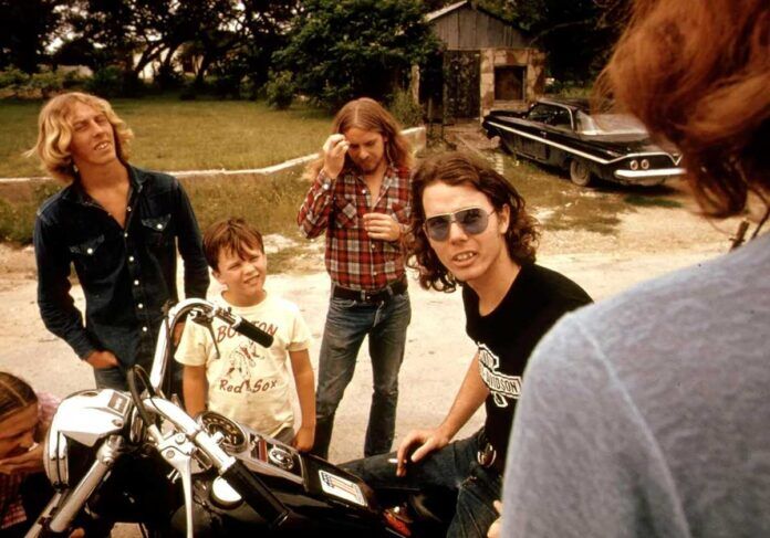 A group of men and a boy stand outdoors near a motorcycle. One man sits on the motorcycle wearing sunglasses. Others stand around, one in a plaid shirt wiping his face. There is grass, trees, and a classic car in the background.