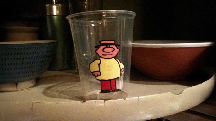 A clear plastic cup features a cartoon character with a hat and round nose, wearing a yellow shirt and red pants. The cup is placed on a slightly worn, curved shelf, with a blue bowl and other items in the background.