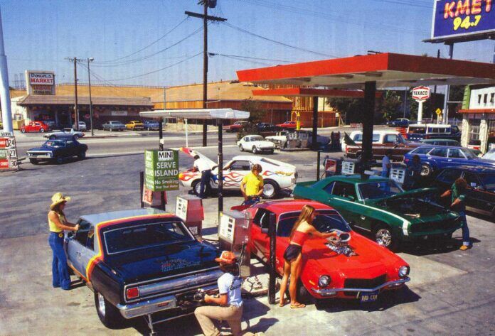 People in 1980s-style clothing pump gas into classic muscle cars at a busy retro gas station. Vintage buildings, parked cars, and vibrant colors add to the nostalgic atmosphere.