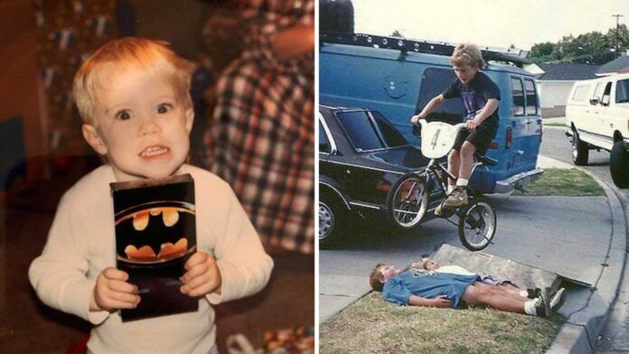 A split image: on the left, a young child with blond hair smiles widely while holding a Batman VHS tape; on the right, a boy jumps his bike over another child lying on a driveway near parked cars.