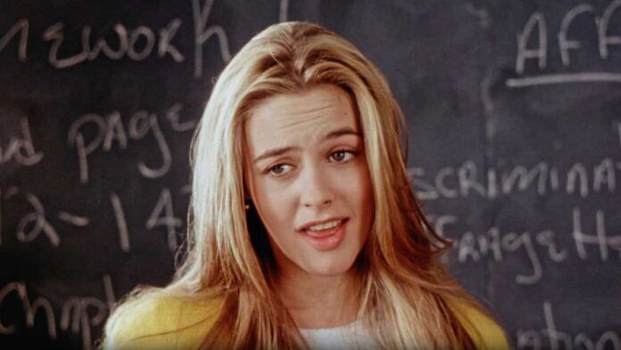 30 movies turn 30 - Alicia Silverstone in Clueless with long blonde hair stands in front of a chalkboard with handwriting on it. They are wearing a yellow jacket and a white top, looking slightly to the side.