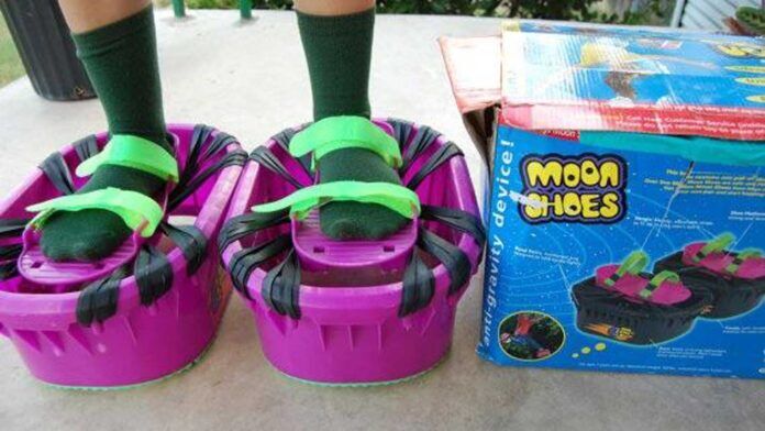 A pair of feet wearing green socks stands in bright purple and green Moon Shoes, which resemble mini-trampolines. A colorful box labeled Moon Shoes is next to them on a concrete surface.